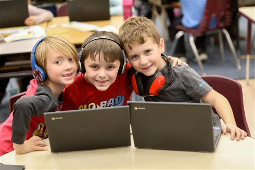 Three elementary-aged students with headphones in front of two Chromebooks 