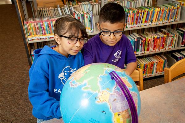 Two elementary students stand at a library table and look at a globe 