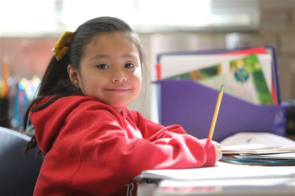 a younger elementary student sits at a desk holding a pencil and smiling 
