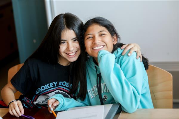 Two middle school students sit with their arms around each other and smile. 