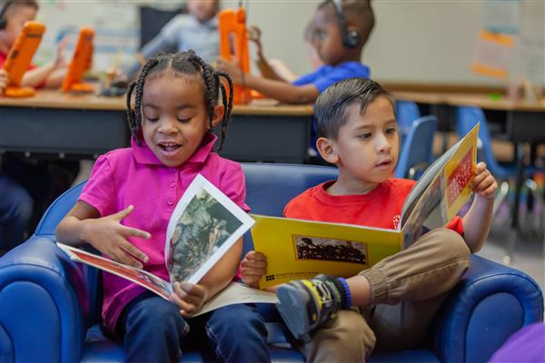Two students sit on a blue chair in their classroom while reading picture books  